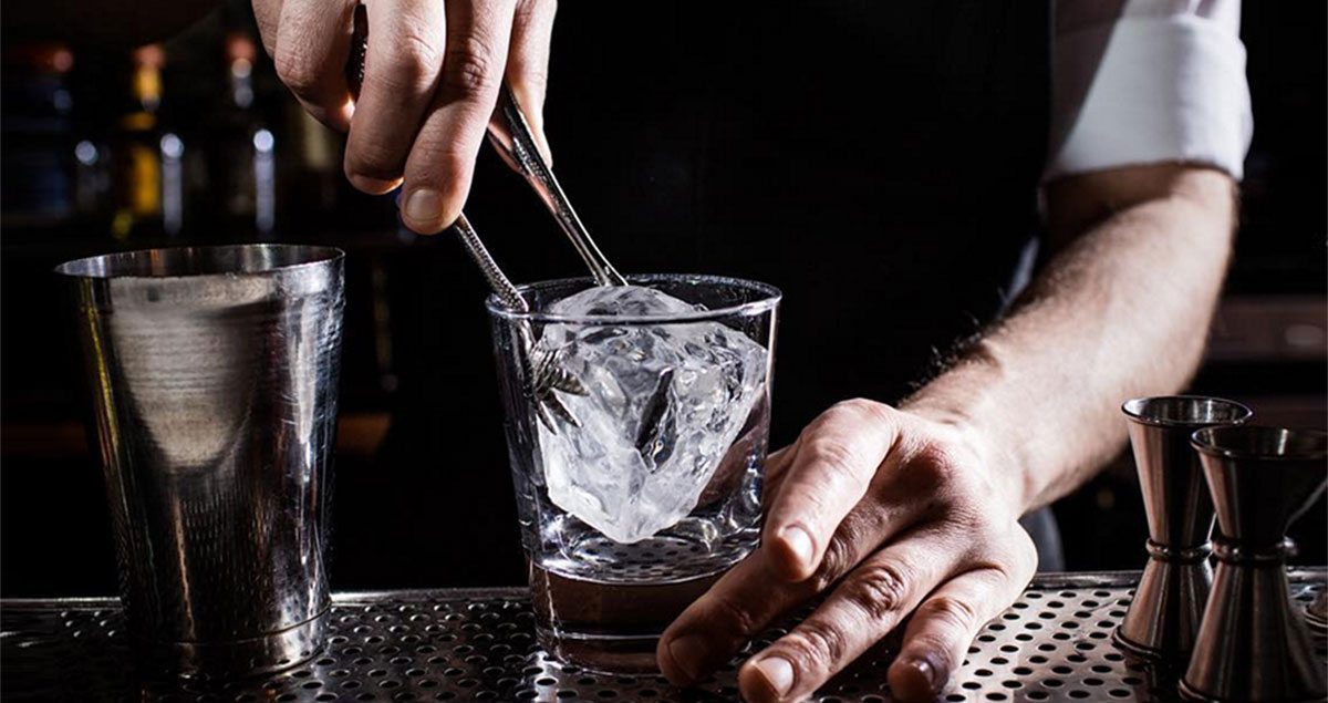 Closeup of a Death and Co. bartender placing a large ice cube into a glass.