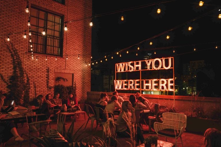 Perspective Shot for the Garden Terrace in the Ramble Hotel. The bar is full of people and there is a red shining billboard showing the message 'Wish you were here'